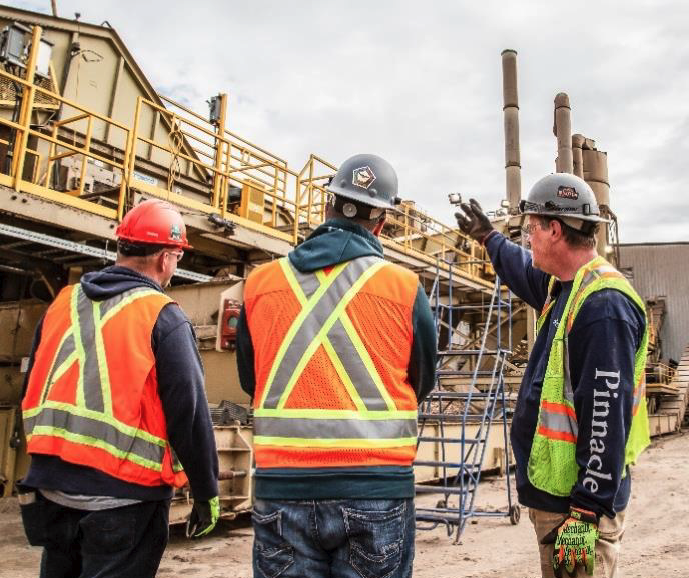 Three workers in safety gear looking at a pellet mill.