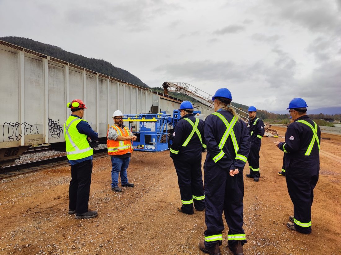 People in safety gear in front of a railcar loading wood pellets.